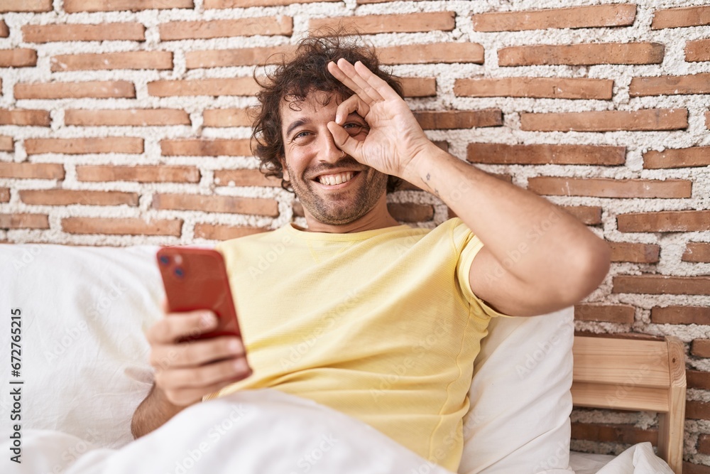 Hispanic young man using smartphone on the bed smiling happy doing ok sign with hand on eye looking through fingers Hispanic young man using smartphone on the bed smiling happy doing ok sign with hand on eye looking through fingers