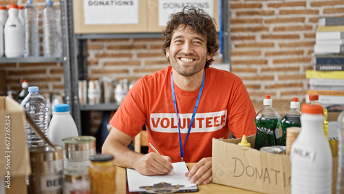 Young hispanic man volunteer writing on clipboard smiling at charity center
