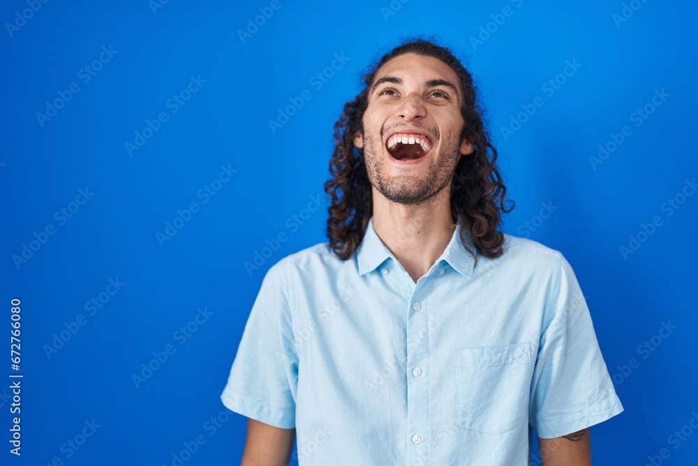 Young hispanic man standing over blue background angry and mad screaming frustrated and furious, shouting with anger. rage and aggressive concept. Young hispanic man standing over blue background angry and mad screaming frustrated and furious, shouting with anger. rage and aggressive concept.