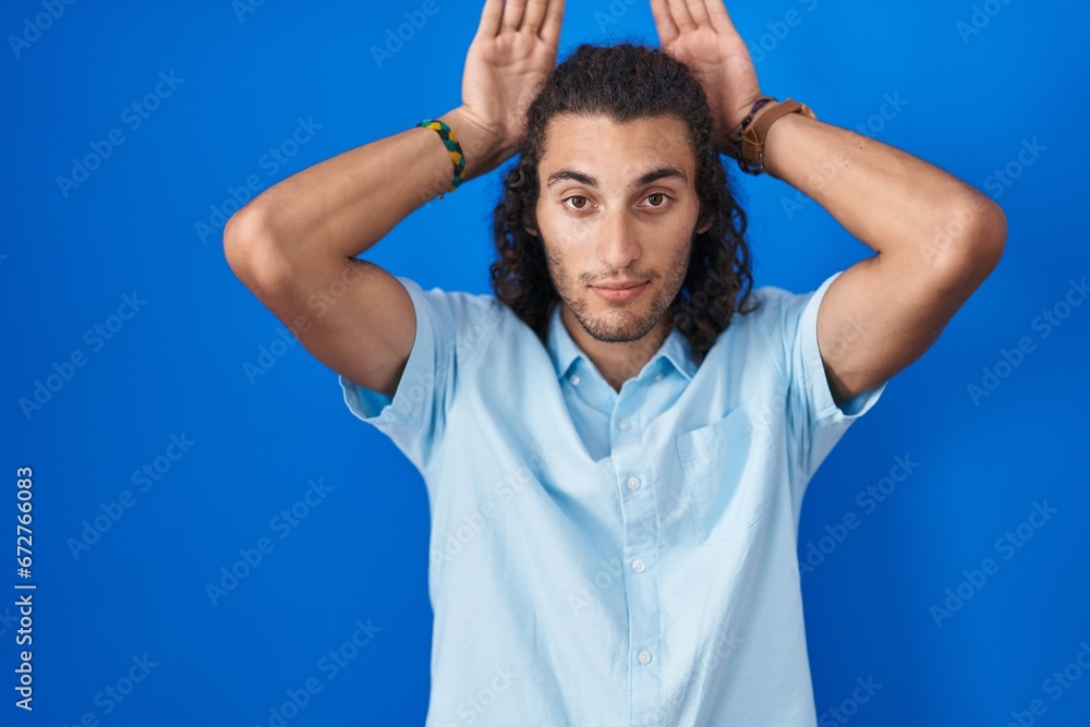 Young hispanic man standing over blue background doing bunny ears gesture with hands palms looking cynical and skeptical. easter rabbit concept. Young hispanic man standing over blue background doing bunny ears gesture with hands palms looking cynical and skeptical. easter rabbit concept.