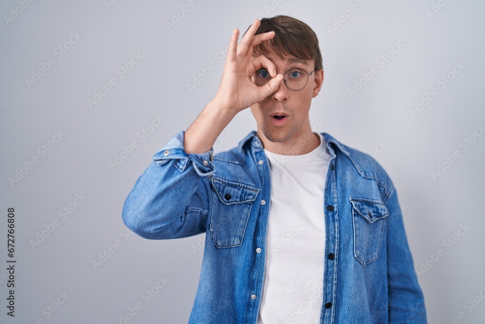 Caucasian blond man standing wearing glasses doing ok gesture shocked with surprised face, eye looking through fingers. unbelieving expression. Caucasian blond man standing wearing glasses doing ok gesture shocked with surprised face, eye looking through fingers. unbelieving expression.