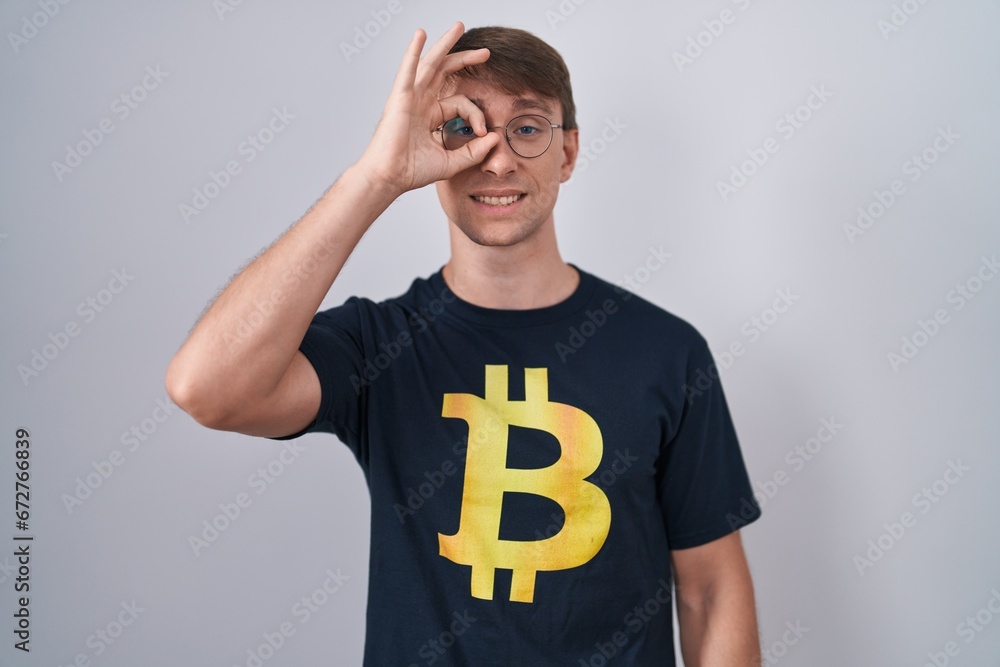 Caucasian blond man wearing bitcoin t shirt doing ok gesture with hand smiling, eye looking through fingers with happy face. Caucasian blond man wearing bitcoin t shirt doing ok gesture with hand smiling, eye looking through fingers with happy face.
