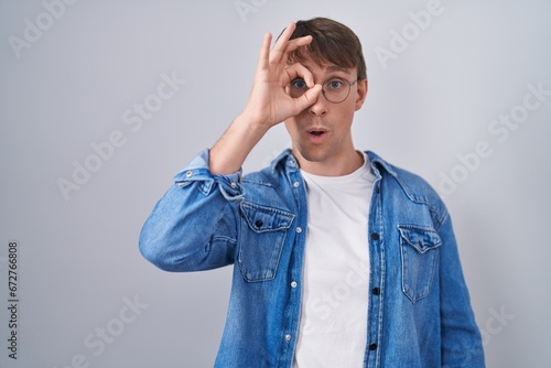 Caucasian blond man standing wearing glasses doing ok gesture shocked with surprised face, eye looking through fingers. unbelieving expression. © Krakenimages.com