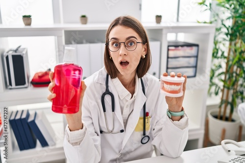 Young caucasian dentist woman holding denture and mouthwash afraid and shocked with surprise and amazed expression  fear and excited face.