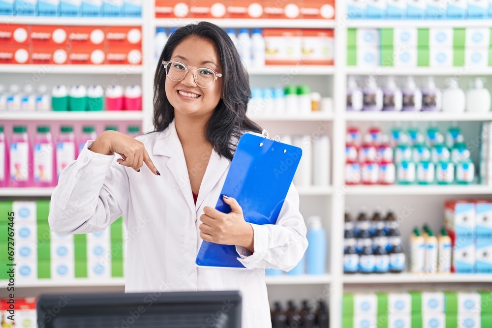 Asian young woman working at pharmacy drugstore holding clipboard pointing finger to one self smiling happy and proud