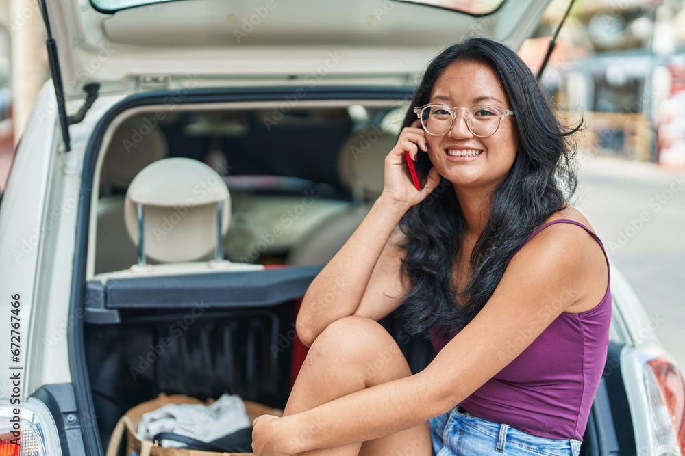 Young chinese woman talking on smartphone sitting on car trunk at street Young chinese woman talking on smartphone sitting on car trunk at street
