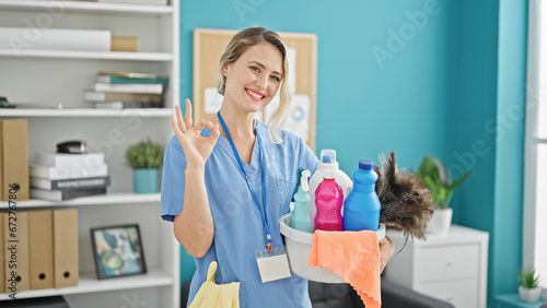 Young blonde woman professional cleaner holding products doing ok gesture at the office