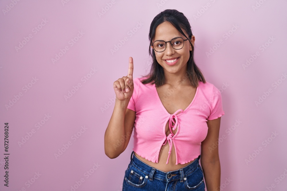 Hispanic young woman standing over pink background wearing glasses showing and pointing up with finger number one while smiling confident and happy. Hispanic young woman standing over pink background wearing glasses showing and pointing up with finger number one while smiling confident and happy.
