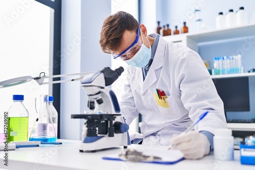 Young man scientist wearing medical mask using microscope writing document at laboratory