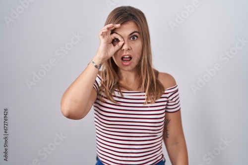Young hispanic woman standing over isolated background doing ok gesture shocked with surprised face, eye looking through fingers. unbelieving expression.