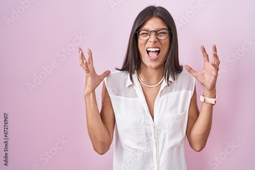 Brunette young woman standing over pink background wearing glasses celebrating mad and crazy for success with arms raised and closed eyes screaming excited. winner concept