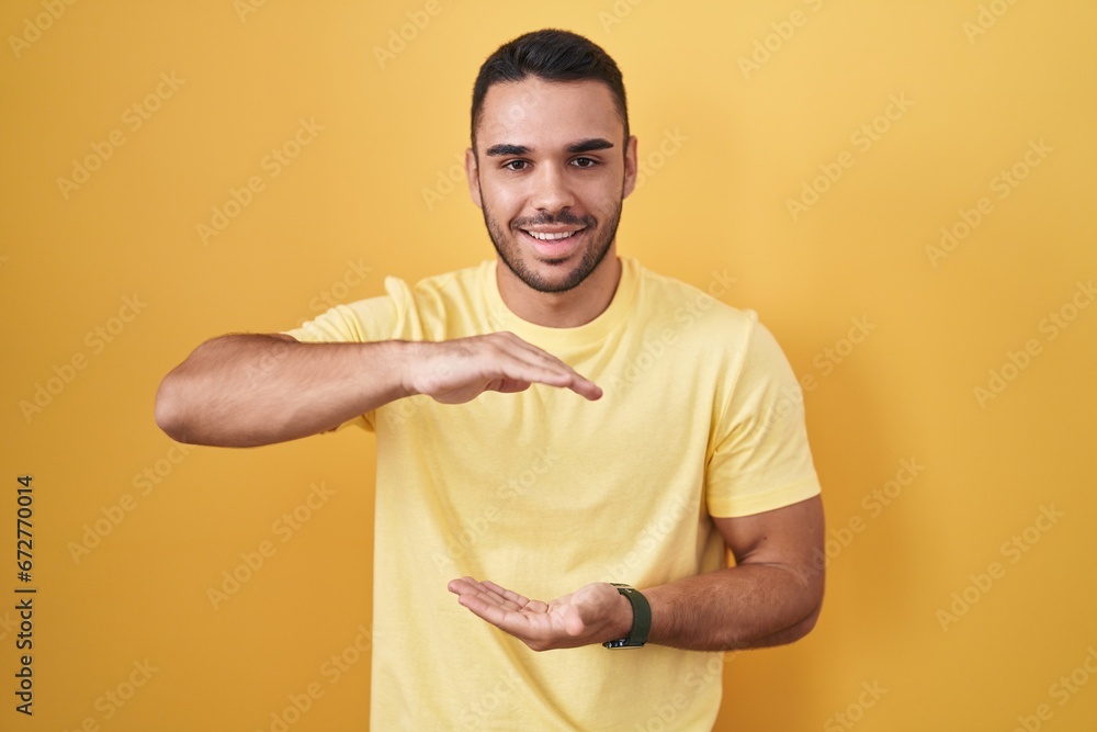 Young hispanic man standing over yellow background gesturing with hands showing big and large size sign, measure symbol. smiling looking at the camera. measuring concept. Young hispanic man standing over yellow background gesturing with hands showing big and large size sign, measure symbol. smiling looking at the camera. measuring concept.