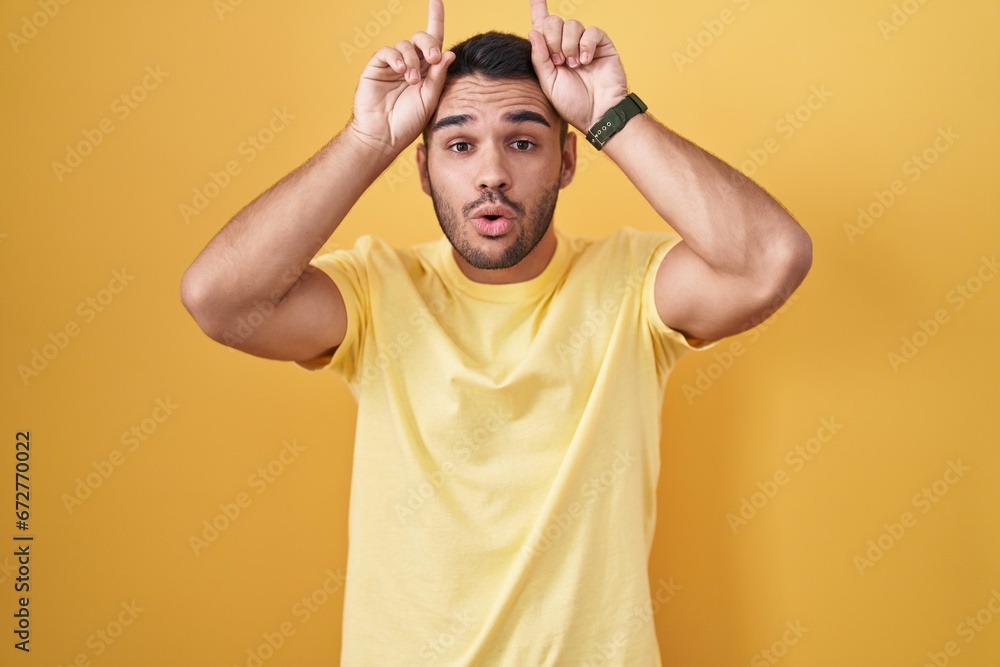 Young hispanic man standing over yellow background doing funny gesture with finger over head as bull horns Young hispanic man standing over yellow background doing funny gesture with finger over head as bull horns