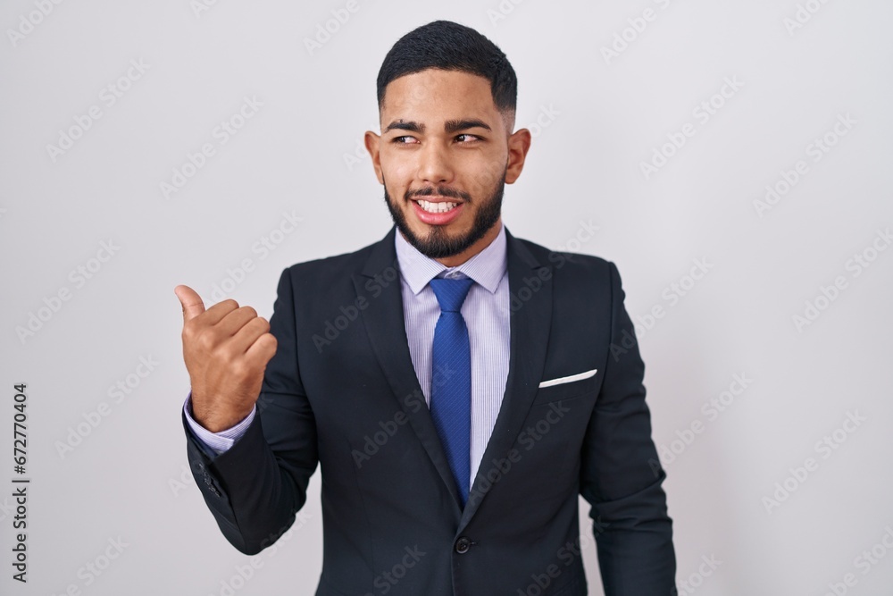 Young hispanic man wearing business suit and tie smiling with happy face looking and pointing to the side with thumb up. Young hispanic man wearing business suit and tie smiling with happy face looking and pointing to the side with thumb up.
