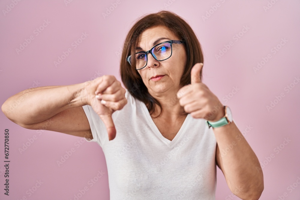 Middle age hispanic woman standing over pink background doing thumbs up and down, disagreement and agreement expression. crazy conflict Middle age hispanic woman standing over pink background doing thumbs up and down, disagreement and agreement expression. crazy conflict