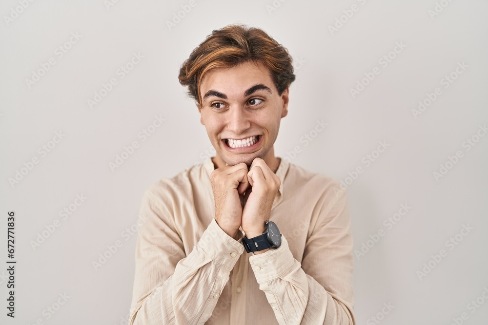 Young man standing over isolated background laughing nervous and excited with hands on chin looking to the side Young man standing over isolated background laughing nervous and excited with hands on chin looking to the side