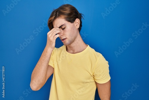 Young man standing over blue background tired rubbing nose and eyes feeling fatigue and headache. stress and frustration concept.