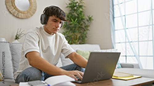 Engrossed young hispanic male student chilling at home, sitting on comfy sofa, focused on studying online via laptop, perfectly relaxed, while seriously soaking up knowledge through headphones.