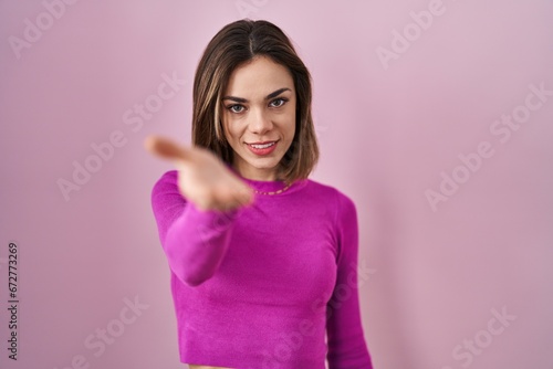 Hispanic woman standing over pink background smiling cheerful offering palm hand giving assistance and acceptance.