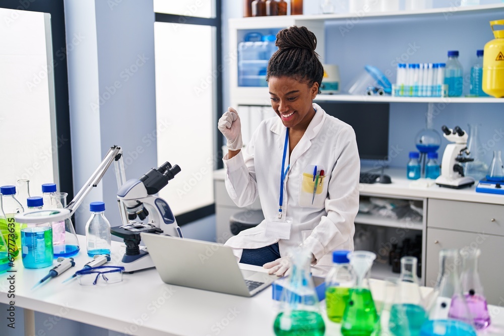 Young african american with braids working at scientist laboratory with laptop screaming proud, celebrating victory and success very excited with raised arm Young african american with braids working at scientist laboratory with laptop screaming proud, celebrating victory and success very excited with raised arm