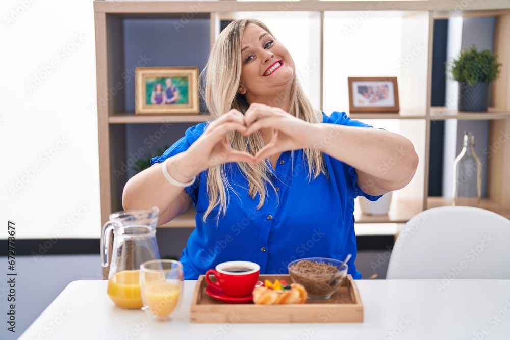 Caucasian plus size woman eating breakfast at home smiling in love doing heart symbol shape with hands. romantic concept.