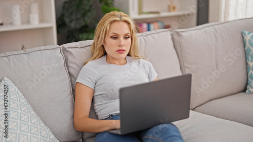 Young blonde woman using laptop sitting on sofa at home