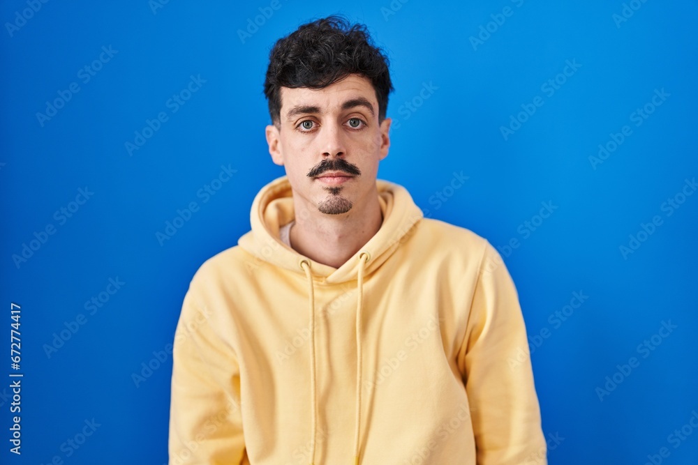 Fototapeta premium Hispanic man standing over blue background relaxed with serious expression on face. simple and natural looking at the camera.