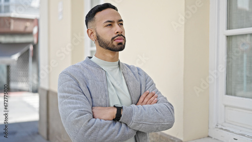 Young hispanic man standing with serious expression and crossed arms at street
