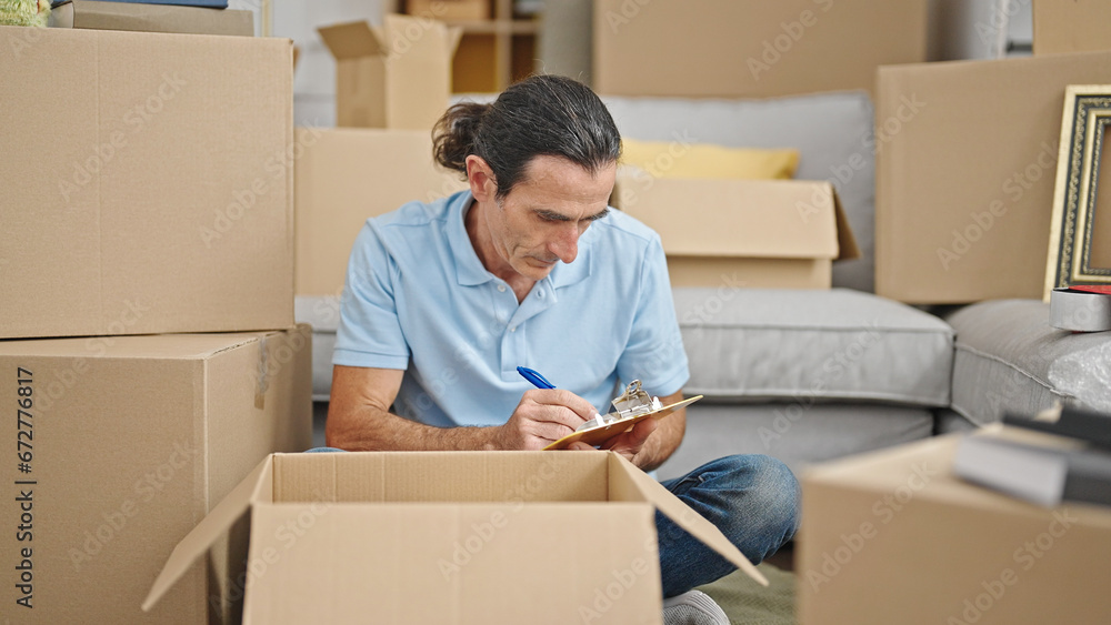 Middle age man checking cardboard box writing on clipboard at new home Middle age man checking cardboard box writing on clipboard at new home