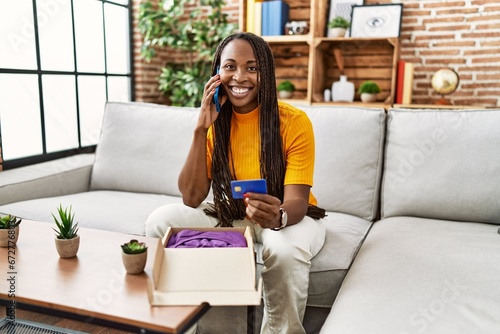 African american woman talking on smartphone holding credit card for online purchase at home
