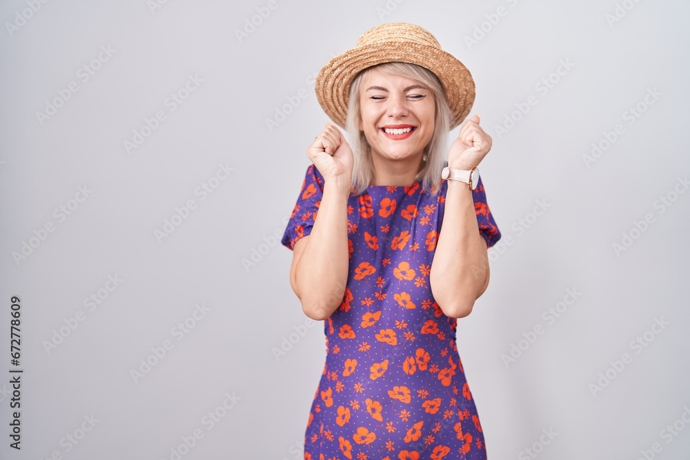 Young caucasian woman wearing flowers dress and summer hat excited for success with arms raised and eyes closed celebrating victory smiling. winner concept.
