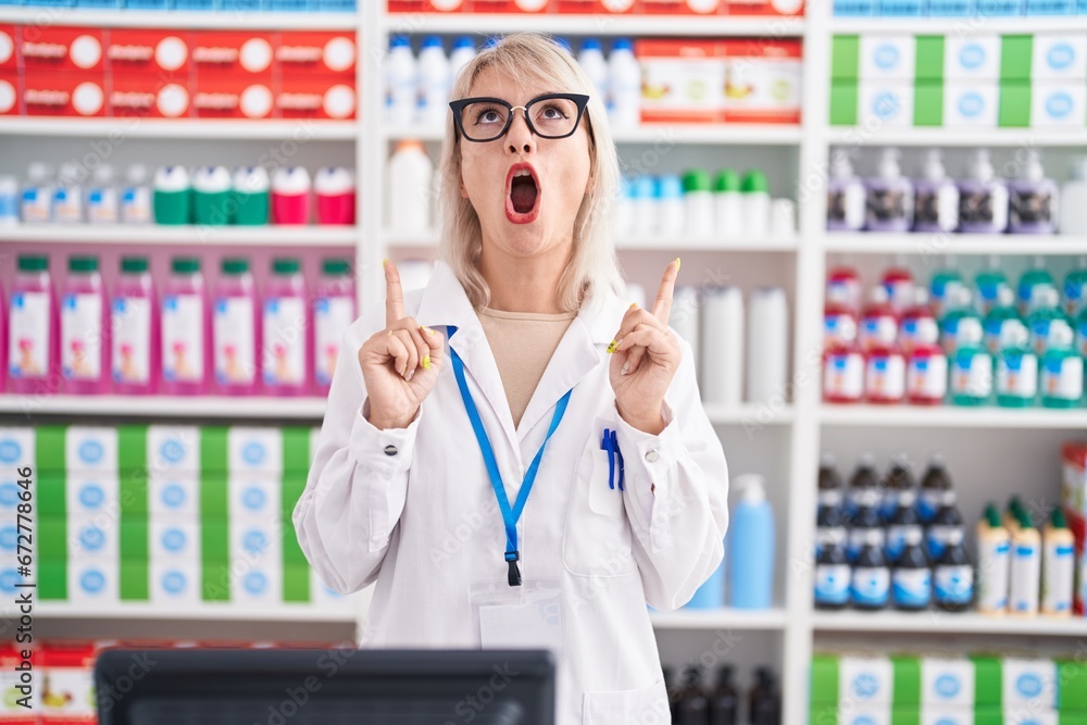 Young caucasian woman working at pharmacy drugstore amazed and surprised looking up and pointing with fingers and raised arms. Young caucasian woman working at pharmacy drugstore amazed and surprised looking up and pointing with fingers and raised arms.