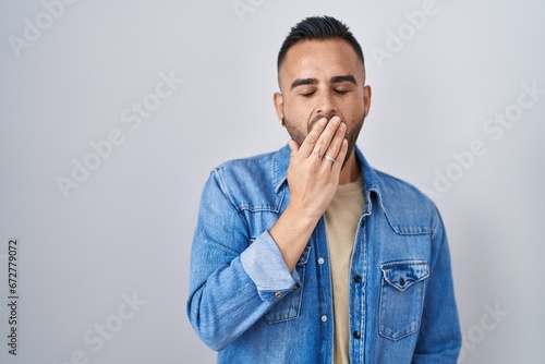 Young hispanic man standing over isolated background bored yawning tired covering mouth with hand. restless and sleepiness.