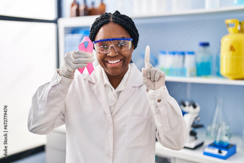 Naklejka premium African american woman working at scientist laboratory holding pink ribbon surprised with an idea or question pointing finger with happy face, number one
