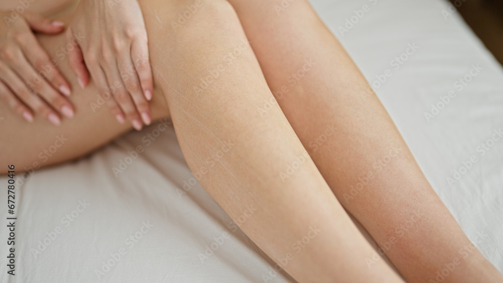 Young redhead woman wearing lingerie applying skin care lotion on leg at bedroom