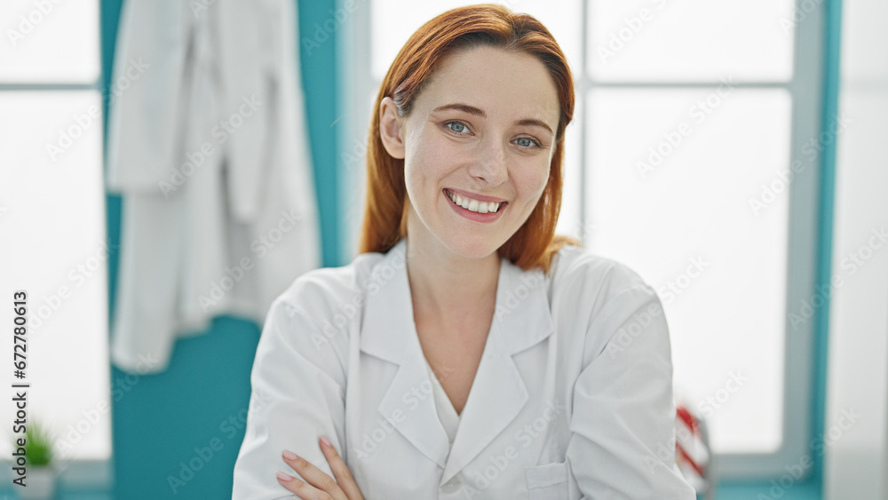 Young redhead woman doctor smiling confident sitting on table with arms crossed gesture at clinic Young redhead woman doctor smiling confident sitting on table with arms crossed gesture at clinic