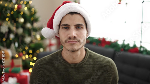 Young hispanic man sitting on sofa by christmas tree with relaxed expression at home