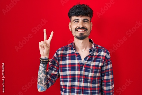Young hispanic man with beard standing over red background showing and pointing up with fingers number two while smiling confident and happy.