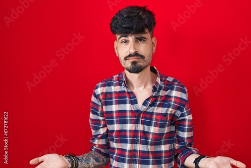 Young hispanic man with beard standing over red background clueless and confused with open arms, no idea concept. © Krakenimages.com