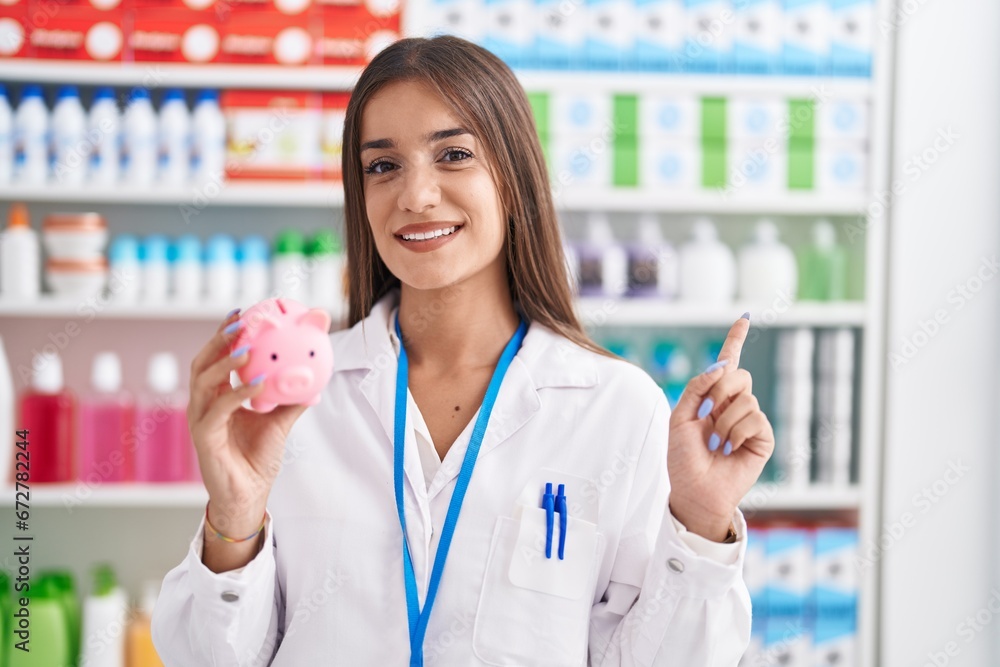 Young brunette woman working at pharmacy drugstore holding piggy bank smiling happy pointing with hand and finger to the side