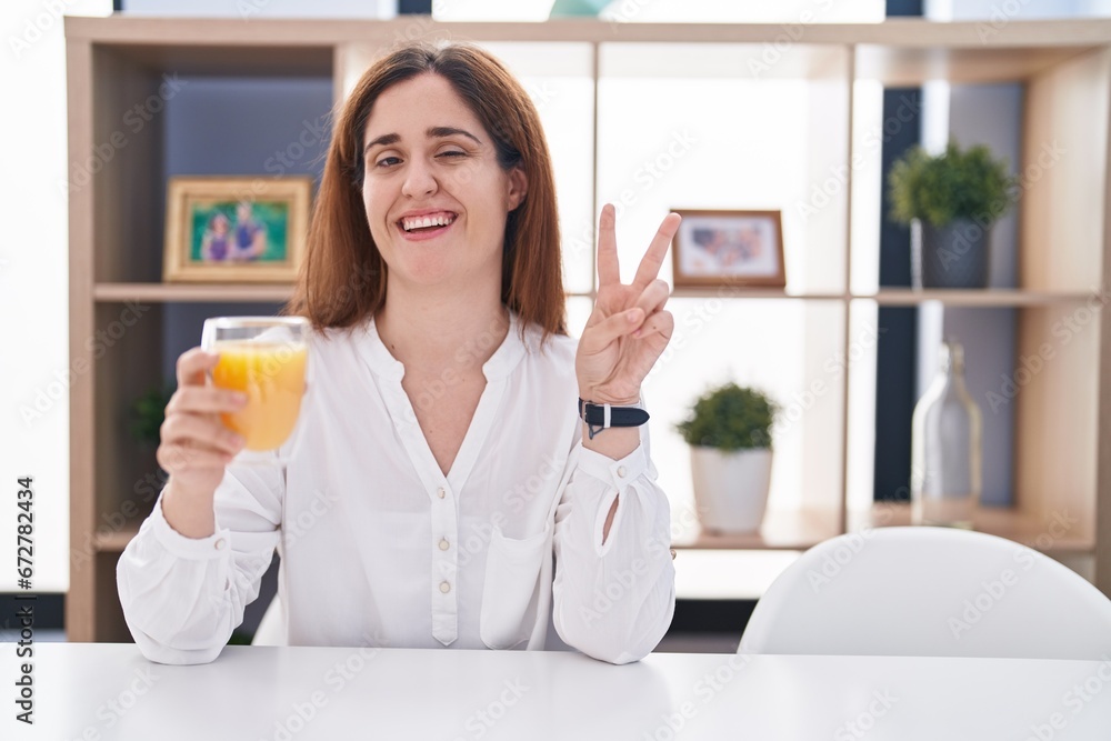 Brunette woman drinking glass of orange juice smiling with happy face winking at the camera doing victory sign with fingers. number two. Brunette woman drinking glass of orange juice smiling with happy face winking at the camera doing victory sign with fingers. number two.