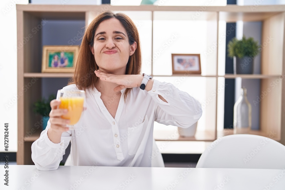 Brunette woman drinking glass of orange juice cutting throat with hand as knife, threaten aggression with furious violence Brunette woman drinking glass of orange juice cutting throat with hand as knife, threaten aggression with furious violence