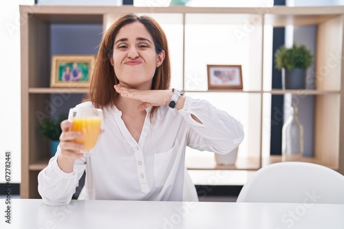 Brunette woman drinking glass of orange juice cutting throat with hand as knife  threaten aggression with furious violence