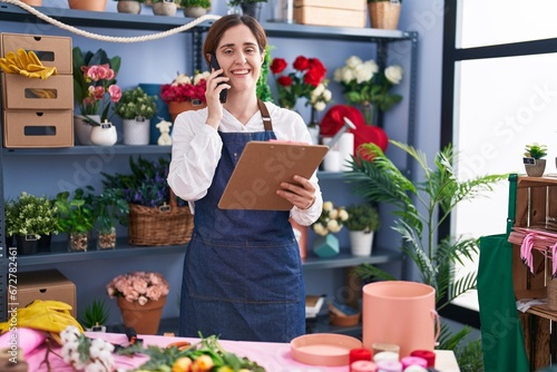 Young woman florist talking on smartphone reading clipboard at florist shop
