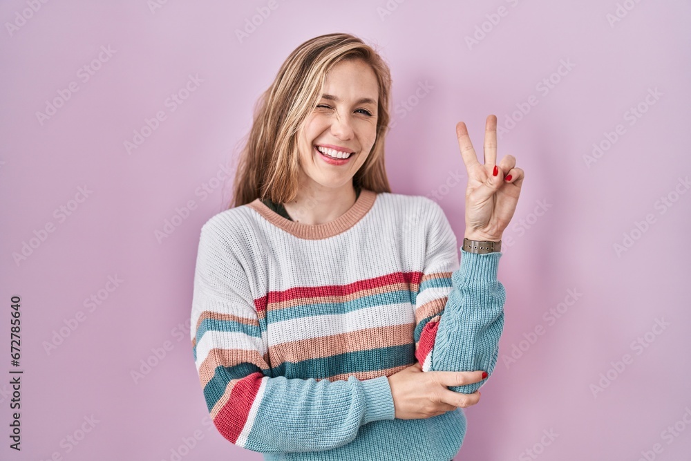 Young blonde woman standing over pink background smiling with happy face winking at the camera doing victory sign with fingers. number two. Young blonde woman standing over pink background smiling with happy face winking at the camera doing victory sign with fingers. number two.
