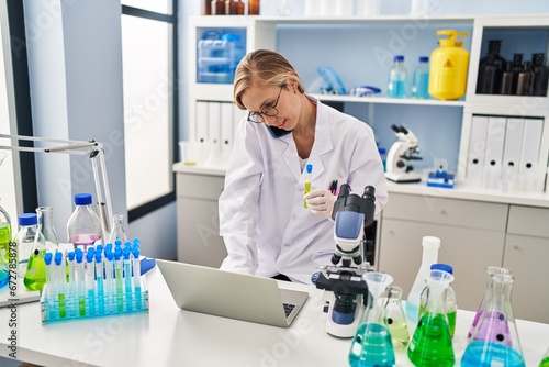 Young blonde woman scientist talking on smartphone holding test tube at laboratory