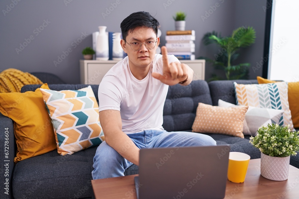 Young asian man using laptop at home sitting on the sofa pointing with finger up and angry expression, showing no gesture