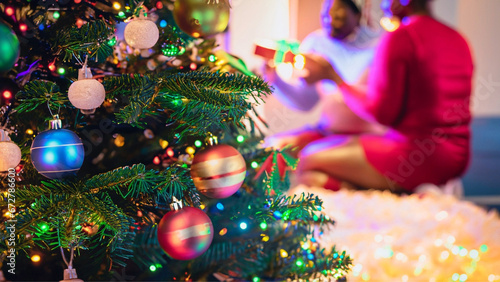 Árvore de Natal bem focada em primeiro plano e pessoas com presentes desfocadas ao fundo.