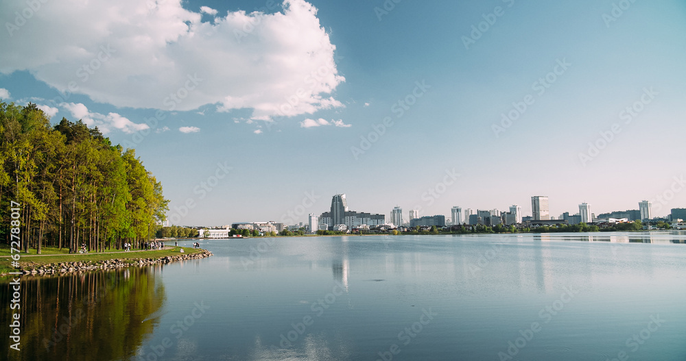 Fototapeta premium Minsk, Belarus. Reservoir Drozdy. View On Forest, Public Park And City On Background. People Resting Walking At Riverside In Summer Day. City Pond And Park. Concept Of Rest And Weekends.