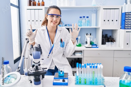 Young hispanic woman working at scientist laboratory showing and pointing up with fingers number seven while smiling confident and happy.